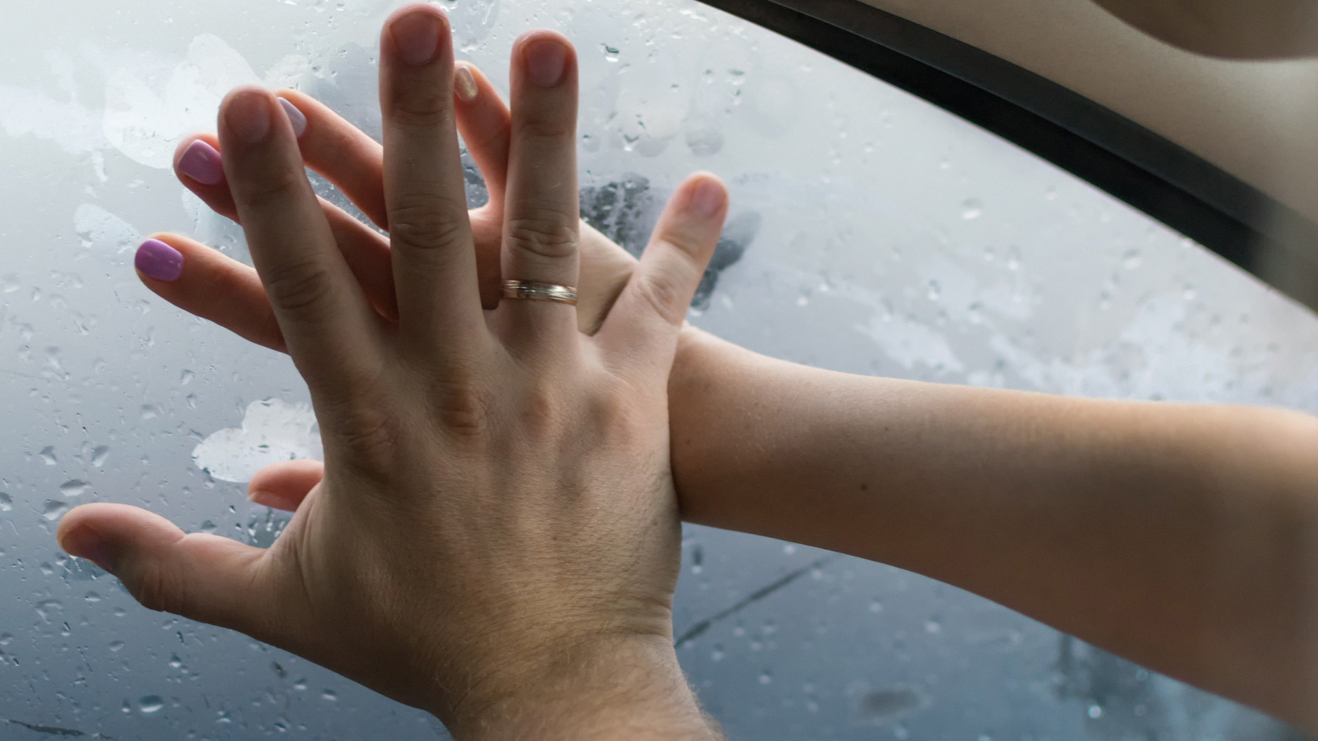 Hands of a man and a woman against a foggy car window. The man's hand has a ring while pushing the woman's against the window